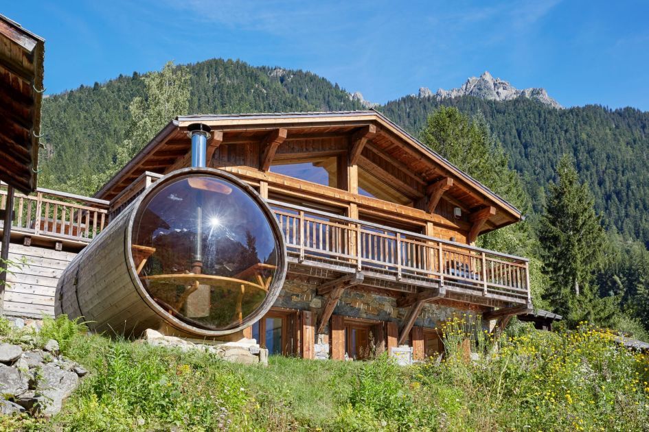 Eco Lodge stands tall in front of tree-topped mountain views. In the foreground, the chalet's superior barrel sauna is positioned. Blue skies paint the picture of a true summer mountain retreat in the Alps.