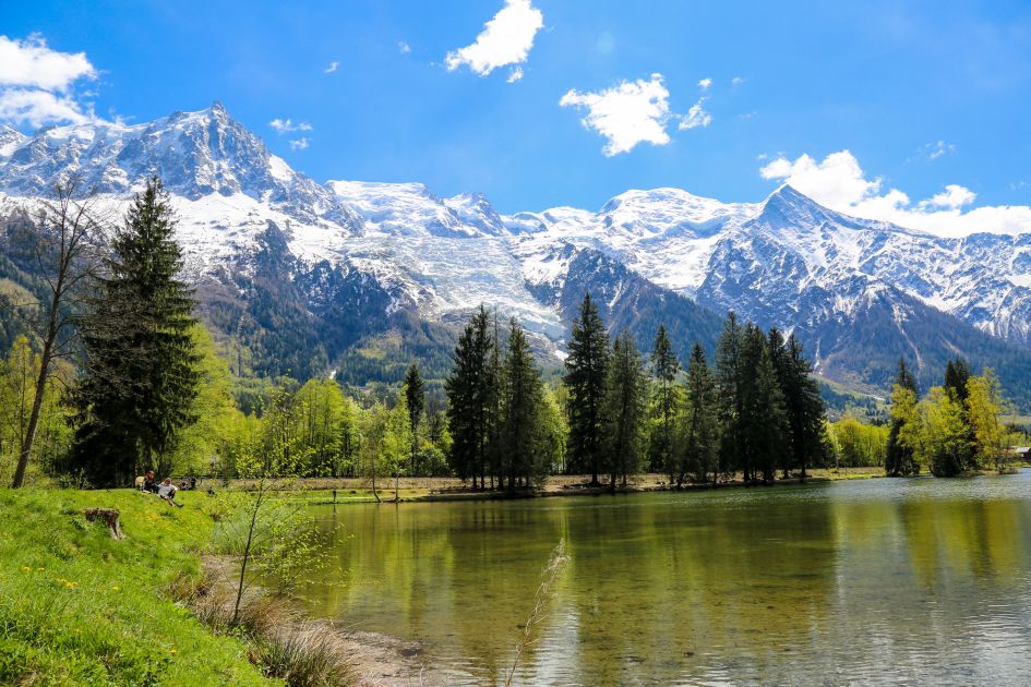 Gaillands Lake in Chamonix features a magnificent mountain backdrop with blues skies, behind the reflective body of water and green trees found at the forefront of the image - a must-visit for your lakes and mountain holiday in Chamonix!