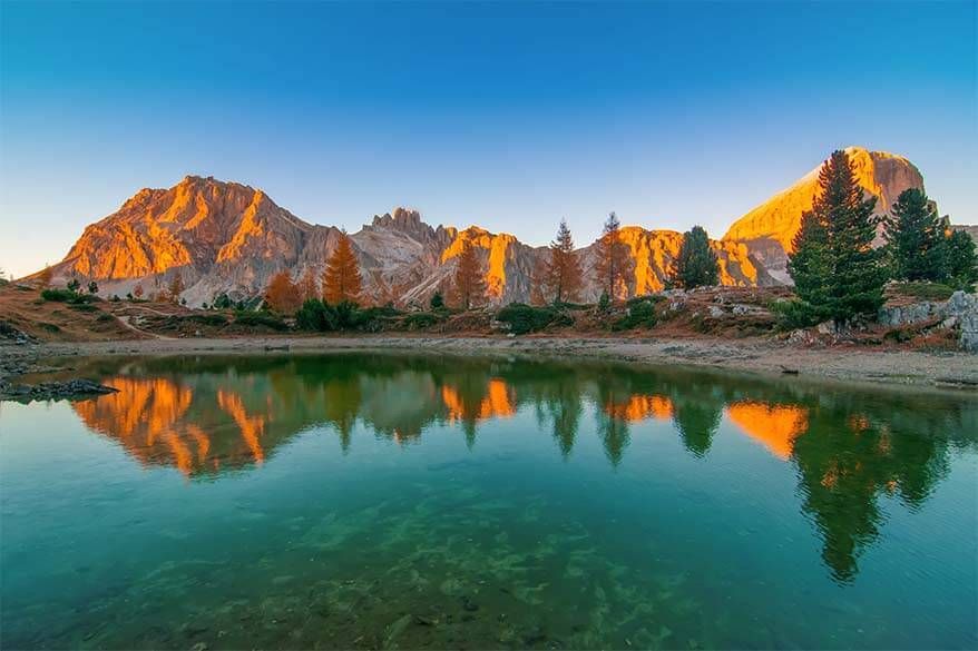 Lago Limides looks beautiful at sunset. A beautiful green body of water mirrors the golden mountains glowing in the sunset in the background.