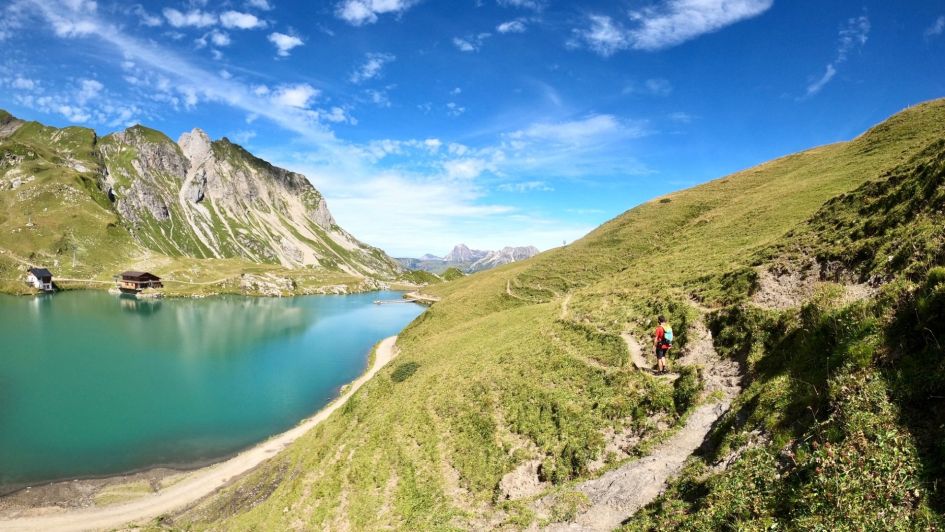 Lake Zürsersee shines under blue skies and sunshine, whilst hikers enjoy a walk around the lake on the green hillside next to the lake.