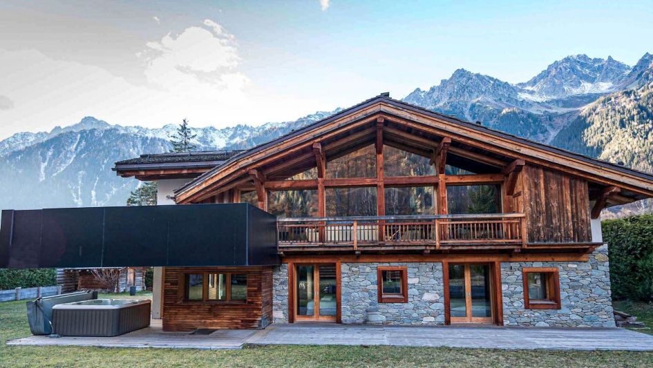 Chalet Bonami's stone and wooden facade is just a taster of what this luxury chalet has inside. There is also an outdoor hot tub seen here, covered by the chalet's large first floor balcony, and mountain views in the background.