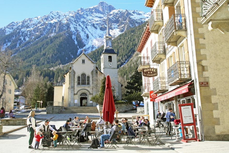 People enjoying a coffee alfresco in 
a cafe in the centre of Chamonix in autumn. The church and immense mountain landscape can be found in the background.