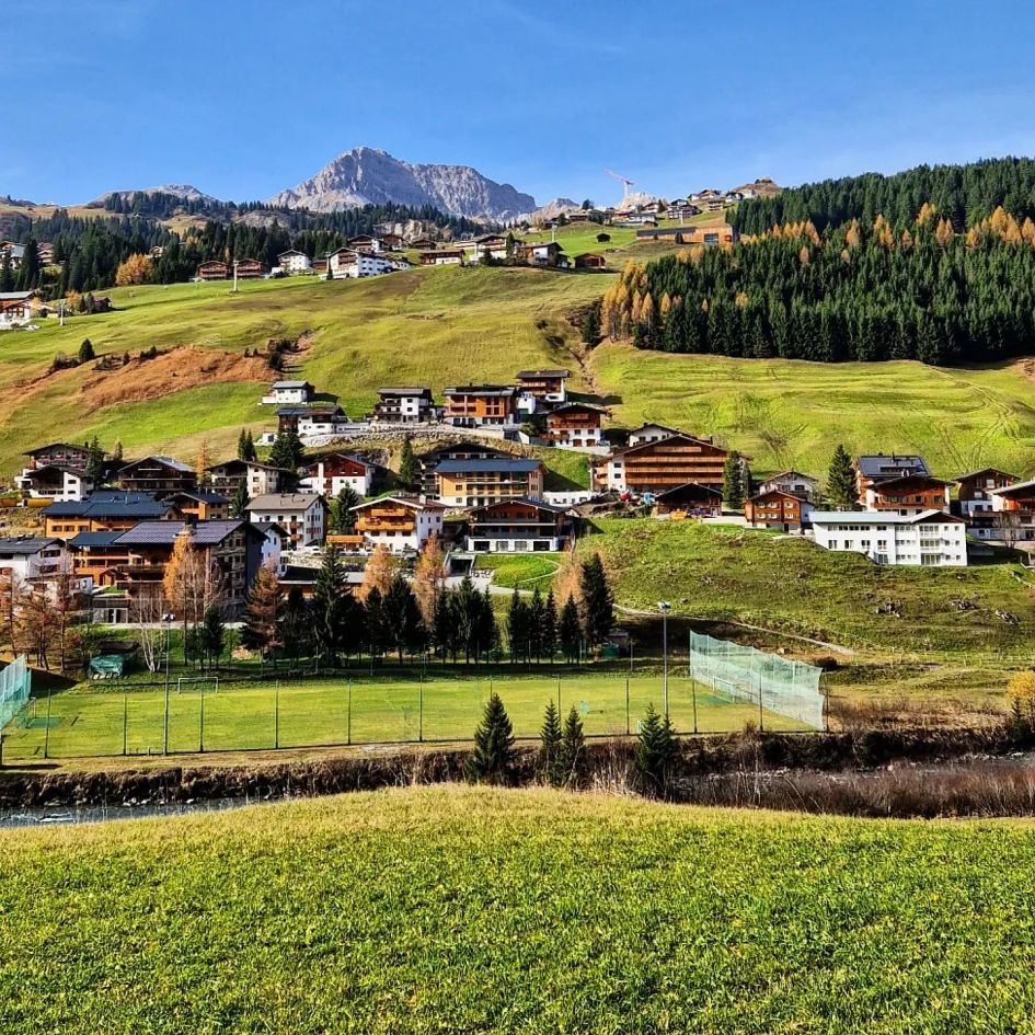 Lech in Autumn is beautiful! A football pitch is found at the foot of the hill. A number of freestanding chalets and apartment buildings ascend above. Higher mountain peaks are seen towards the back of this image, protruding over the landscape and cutting through the blue skies.