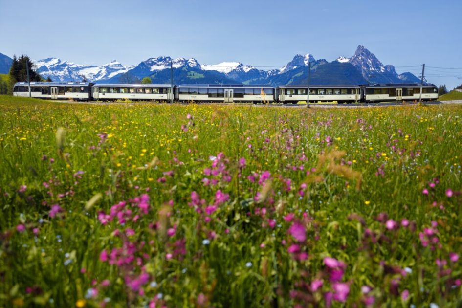 Image of the Montreux Oberland Bernois Railway (MOB) crossing between fields and a mountain view in Gstaad in the Summer. Some incredible views to have on a Gstaad Summer holiday.