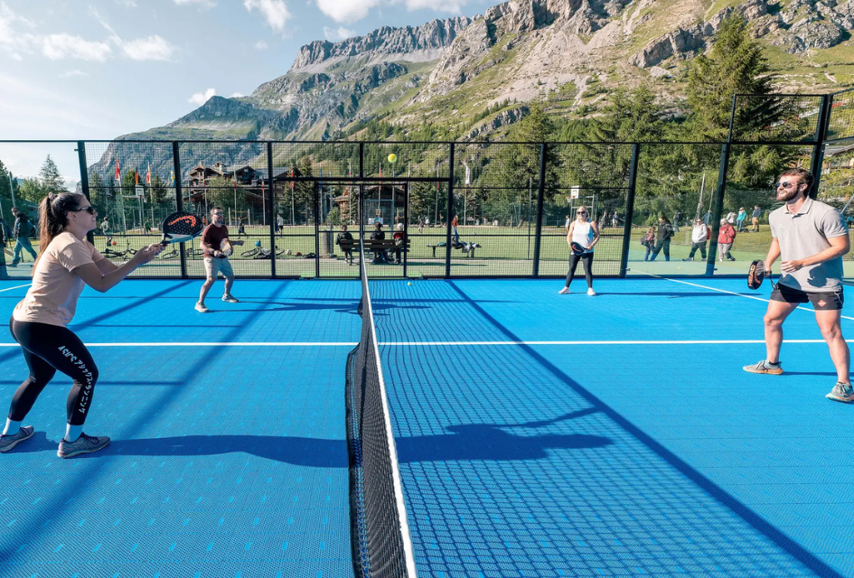A group of 4 playing padel in Val d'Isere with the ball in the air. The game is being played before stunning mountain scenery making this one of the best places to play padel in the Alps. 